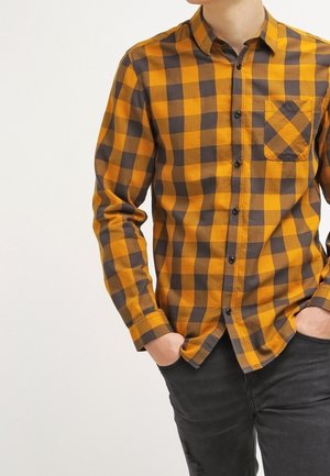 Mężczyzna ubrany w pomarańczowo-czarną kratowaną koszulę na guziki, z rękami w kieszeniach, w zestawieniu z ciemnymi spodniami na jasnym tle.