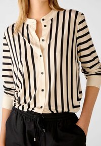 Černobílá pruhovaná halenka s kulatým výstřihem, zapínáním na knoflíky vpředu a dlouhými rukávy. Hladká látka s kontrastními černými knoflíky.
