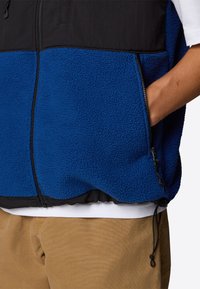 Niebieska kamizelka polarowa z czarną górną częścią, z zapięciem na zamek błyskawiczny z przodu i kieszeniami po bokach. Posiada ściągacz na dolnym wykończeniu oraz teksturowaną tkaninę.