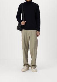 Person som bär svart turtleneck, beige cargo-byxor, vita sneakers och har en liten svart korsbodyväska mot en enfärgad bakgrund.