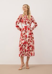 Bloemenprint jurk met lange mouwen, met rode en roze bloemen op een lichte achtergrond. A-lijnvorm en geplooide textuur. Gouden sandalen met bandjes.