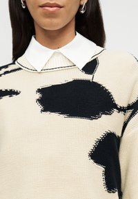 Κοντινό πλάνο ατόμου που φοράει εκρού πλεκτό πουλόβερ με μεγάλα μαύρα αφηρημένα σημεία και λευκό πουκάμισο με γιακά από κάτω, ορατά ασημένια σκουλαρίκια.