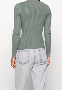 Vrouw in een nauwsluitende geribbelde lange mouwen top in gedempt groen, gecombineerd met hoge taille lichtgrijze gewassen jeans, gezien vanuit de achterkant.