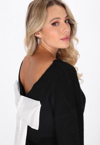 Schwarzer Strickpullover mit tiefem V-Ausschnitt am Rücken und einer großen weißen Stoffschleife hinten. Die Textur wirkt weich und gemütlich.