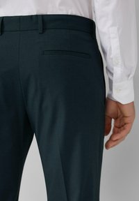 Pantalons ajustés bleu sarcelle foncé avec une texture lisse, dotés d'une poche arrière et de passants de ceinture simples, assortis à une chemise blanche à manches longues.