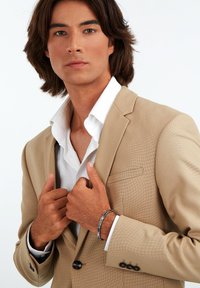 Giacca da abito beige e strutturata con una camicia bianca. Il modello indossa un braccialetto argento e blu. Le mani sono posizionate sul colletto.