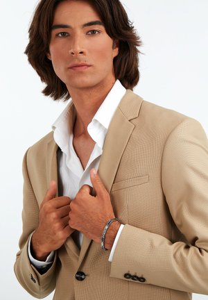 Beige, strukturierte Sakkojacke mit einem weißen Hemd. Das Modell trägt ein silbernes und blaues Armband. Die Hände sind am Kragen positioniert.