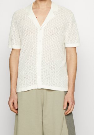 Mężczyzna ubrany w białą dzianinową koszulę na guziki z krótkim rękawem i wzorem w diamenty oraz beżowe spodnie o szerokich nogawkach.