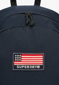 Mochila de tela color navy con una superficie texturizada, cierre doble con tiradores negros y un parche que muestra una bandera americana y "SUPERDRY®".