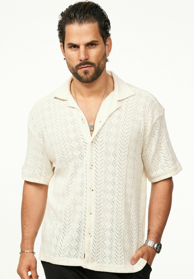 Strickshirt mit kurzen Ärmeln in Creme, mit einem Zickzackmuster und Knopfverschluss. Glatte Textur mit lockerer Passform und offenem Kragen.