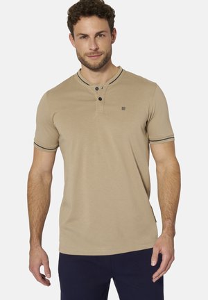 Mann trägt beige kurzärmeliges Henley-Shirt mit zwei Knöpfen und marineblauen Hosen, steht vor einem einfachen hellen Hintergrund.
