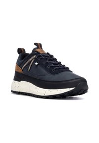 Zapatilla deportiva en azul marino y negro con tela texturizada, detalles en cuero marrón y una media suela blanca moteada. Presenta una suela gruesa y resistente.