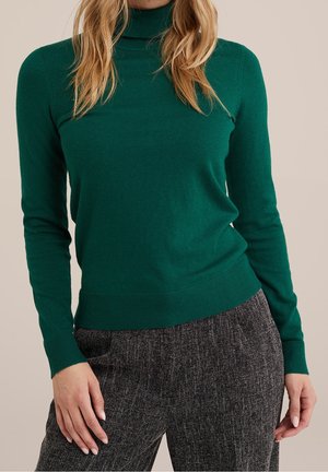 Frau trägt einen engen dunkelgrünen Rollkragenpullover und hoch taillierte graue strukturierte Hosen, steht mit entspannten Händen an den Seiten.