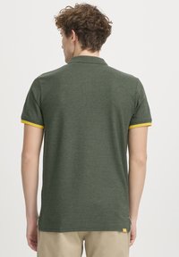 Mørkegrøn polo-shirt med korte ærmer, gule detaljer på kraven og et struktureret stof. Synlig mærkeetiket i kanten.