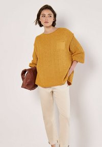Senfgelb gestrickter Pullover mit übergroßem Schnitt und kurzen Ärmeln, mit einer vorderen Tasche. Kombiniert mit hellbeigen cropped Hosen.