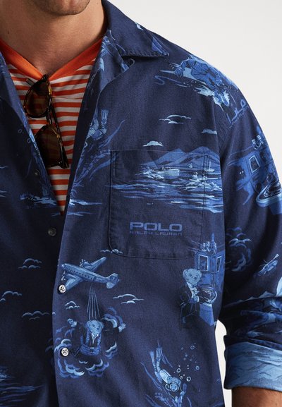 Σκούρο μπλε πουκάμισο με ανοιχτό μπλε vintage σχέδια αεροναυτικής και υποβρύχιας θεματολογίας, λογότυπο "Polo Ralph Lauren" στην τσέπη του στήθους, φορεμένο πάνω από πορτοκαλί ριγέ μπλουζάκι.