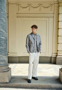 Veste légère grise avec un col, des détails à boutons, et une chemise à rayures en dessous, associées à un pantalon blanc et des chaussures noires.