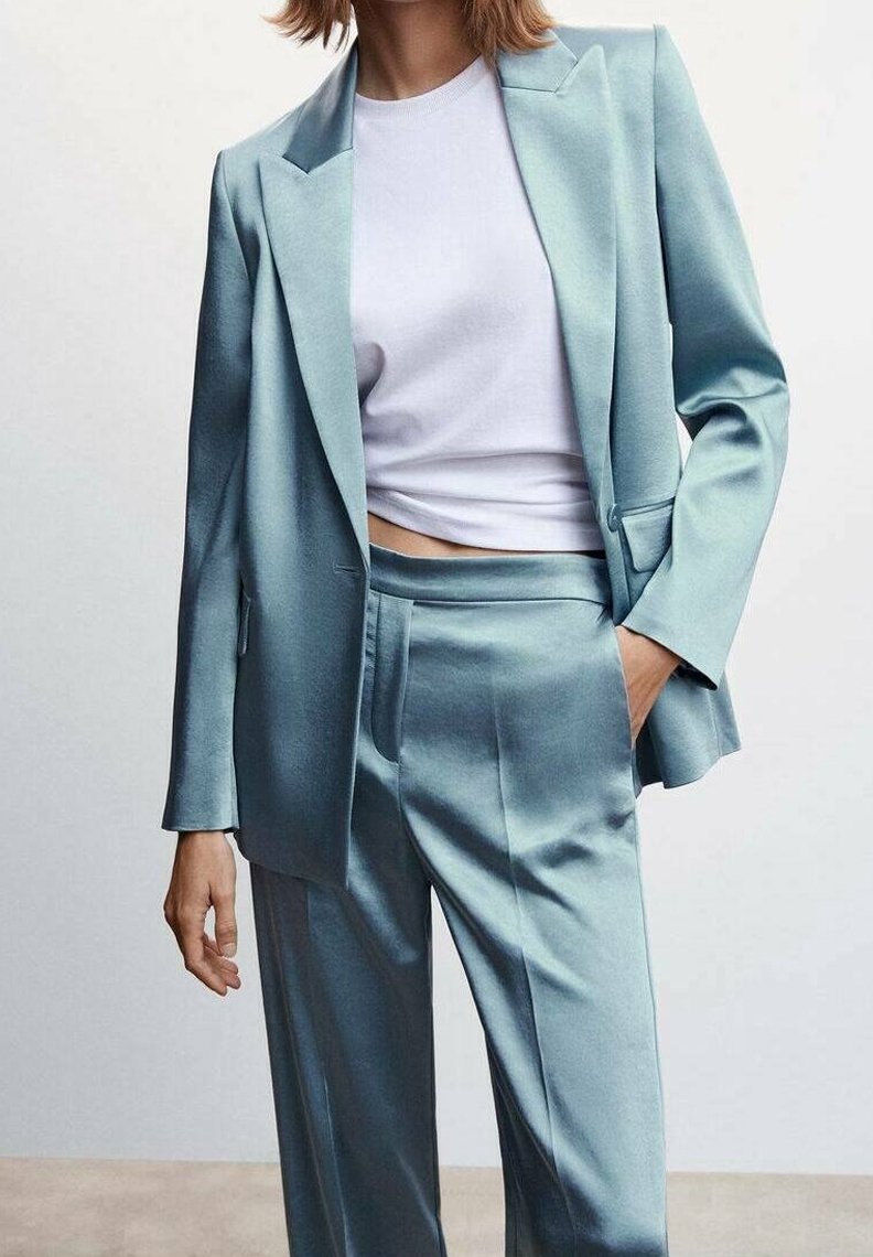 Un costume en satin bleu clair comprenant un blazer ajusté avec des revers crantés et un pantalon droit, associé à un haut blanc ajusté.