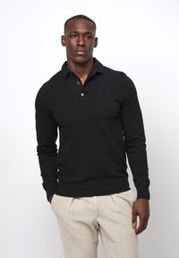 Schwarzer langärmeliger Poloshirt-Pullover mit kragen, zwei vorderen Knöpfen und gerippten Bündchen, kombiniert mit beigen Cordhosen.