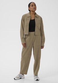 Chaqueta recortada color caqui con grandes bolsillos frontales, combinada con pantalones holgados a juego. Top ajustado negro y zapatillas blancas y grises. Textura suave.