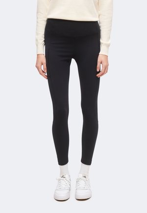 Person iført sorte tætsiddende leggings, hvide ankelsokker, hvide sneakers og en cremefarvet langærmet top, stående mod en ensfarvet baggrund.