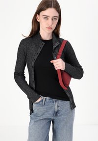 Svart teksturert cardigan med knapper, brukt over en svart skjorte. Parret med lyseblå denimjeans og en rød håndveske med stropp.