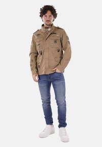 Giacca militare color tan con toppe; presenta una chiusura lampo frontale, quattro tasche e un colletto. Indossata sopra una camicia leggera e jeans blu.