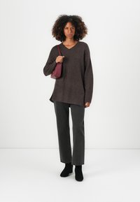Maglione oversize marrone con scollo a V e maniche lunghe, abbinato a jeans dritti grigio scuro e stivali neri alla caviglia. Tiene in mano una borsa a tracolla rossa.