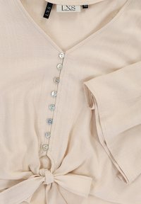 Beige blouse met een V-hals, zachte textuur, acht decoratieve knopen aan de voorkant en een geknoopte details op de taille.