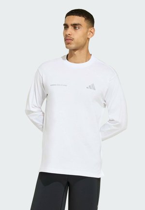 Homme aux cheveux courts portant une chemise blanche à manches longues avec le texte « Running State of Mind » et un pantalon noir, debout les mains dans le dos.