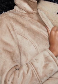 Tan faux-suede jacket met een hoge kraag, zachte textuur en gestikte details. Open geritst om een crèmekleurige fleece voering te onthullen.