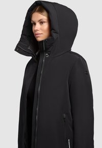 Zwarte geïsoleerde jas met een grote capuchon, ritssluiting en zijzakken. Gladde textuur en minimalistisch ontwerp zonder zichtbare patronen.