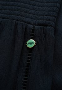 Marineblå stof med en struktureret, plisseret design. Fremhæver en rund, mintgrøn "Superdry" logo knap med guldfarvede accenter nær sømmen.