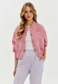 Różowa bawełniana kurtka z kwiatowym haftem, z zatrzaskami i ściągaczami, w połączeniu z lawendowymi spodniami w białą kratkę.