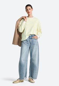 Gestreifter übergroßer Pullover in Gelb und Weiß, hellblaue Jeans mit weitem Bein und ausgefransten Säumen sowie beigen Slippern, kombiniert mit einem braunen Mantel.