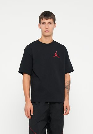 T-shirt in cotone nero con maniche corte, caratterizzato da un logo rosso di Jordan sul petto. La vestibilità è rilassata, mostrando un design semplice.