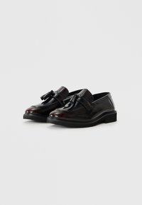 Mocasines de charol negro con punta redondeada, que cuentan con una hebilla decorativa y una suela baja y resistente. Textura suave con un acabado pulido.
