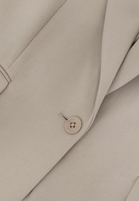 Beige blazerstof med enkelt knap lukket, der viser kraveflip og en del af en lomme.