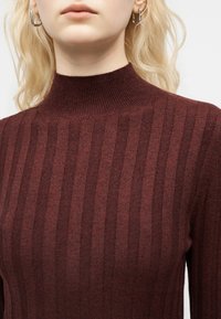 Personne blonde portant des boucles d'oreilles argentées en forme d'anneau et un pull à col roulé côtelé rouge foncé, visible des épaules jusqu'aux lèvres.