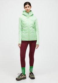Light green puffer jacket with a chevron pattern, hood, and full zipper. Paired with burgundy leggings and multi-colored hiking shoes.