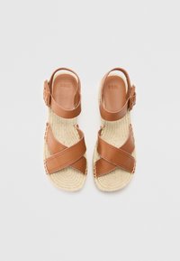 Sandales en cuir marron avec un design à lanières croisées, semelles en jute tressé, boucle ajustable à la cheville et texture lisse. Logo de la marque embossé à l'intérieur.