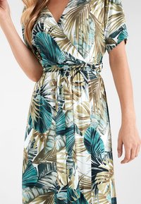 Robes portefeuille florales avec des motifs de feuilles tropicales vertes, bleues et dorées. Tissu doux, manches courtes et design cintré à la taille.