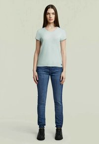 Sky blue majica s kratkimi rokavi, okroglim ovratnikom, v kombinaciji z temno modrimi skinny kavbojkami in črnimi čevlji, ki stoji ob nevtralni ozadju.