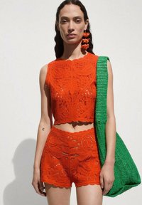 Häkeloberteil und Shorts-Set in lebhaftem Orange mit komplexen Mustern; kombiniert mit einer strukturierten grünen, gewebten Schultertasche.