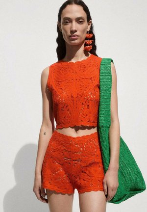 Häkeloberteil und Shorts-Set in lebhaftem Orange mit komplexen Mustern; kombiniert mit einer strukturierten grünen, gewebten Schultertasche.