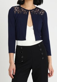 Mörkblå kort cardigan med spetsdetaljer vid axlarna, rund halsringning och trekvartslånga ärmar, kombinerad med högmidjade svarta byxor.