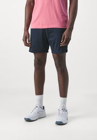 Námořnické sportovní šortky s hladkým povrchem; v kombinaci s bílými ponožkami a bílými teniskami s modrými akcenty. Model má na sobě růžové tričko.