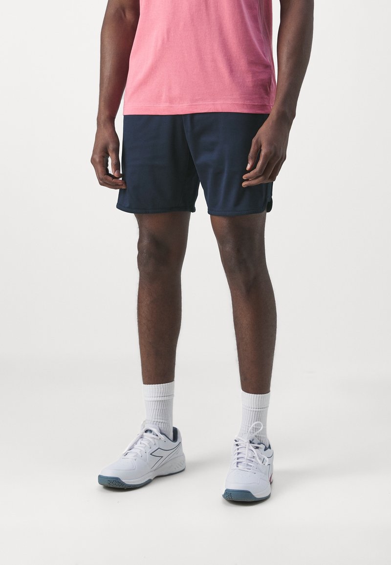 Námořnické sportovní šortky s hladkým povrchem; v kombinaci s bílými ponožkami a bílými teniskami s modrými akcenty. Model má na sobě růžové tričko.