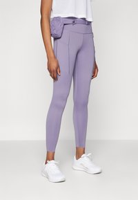 Leggings violets en tissu extensible, avec des poches latérales et une pochette à la taille assortie. Accompagnés de chaussures de sport blanches.