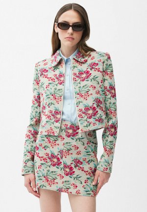 Frau mit Sonnenbrille, hellblauem Hemd zum Knöpfen und einem geblümten Jackett sowie einem passenden Rock in Rot-, Pink- und Grüntönen.